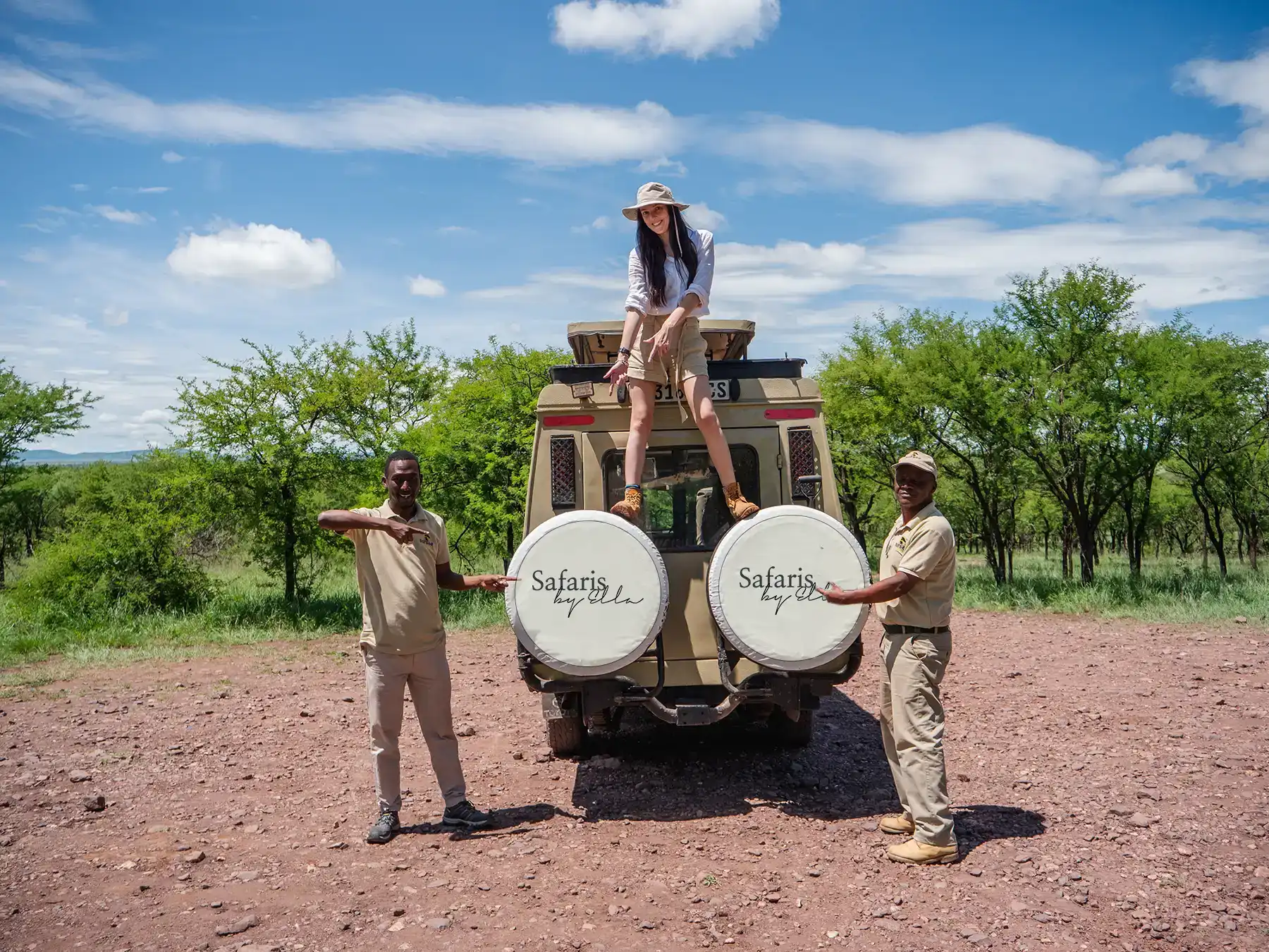 Ella McKendrick sitting on top of a Safaris by Ella branded safari vehicle with two local tour operator guides standing beside it in the Serengeti, Tanzania