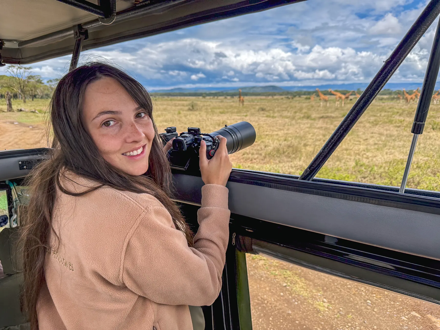 Ella McKendrick smiling from inside a safari vehicle holding a camera with giraffes on the plains behind her on safari in Kenya