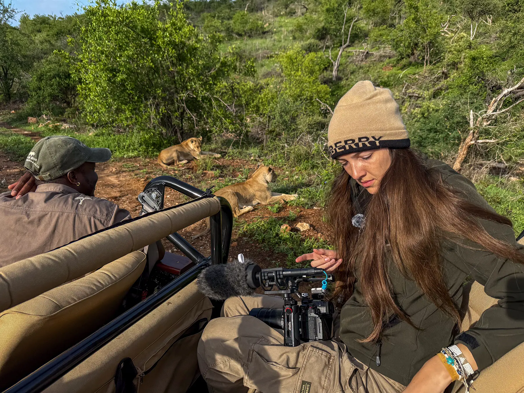Ella McKendrick adjusting a video camera as two lionesses rest on the hillside just behind the safari vehicle on safari in Olifants West Game Reserve, South Africa