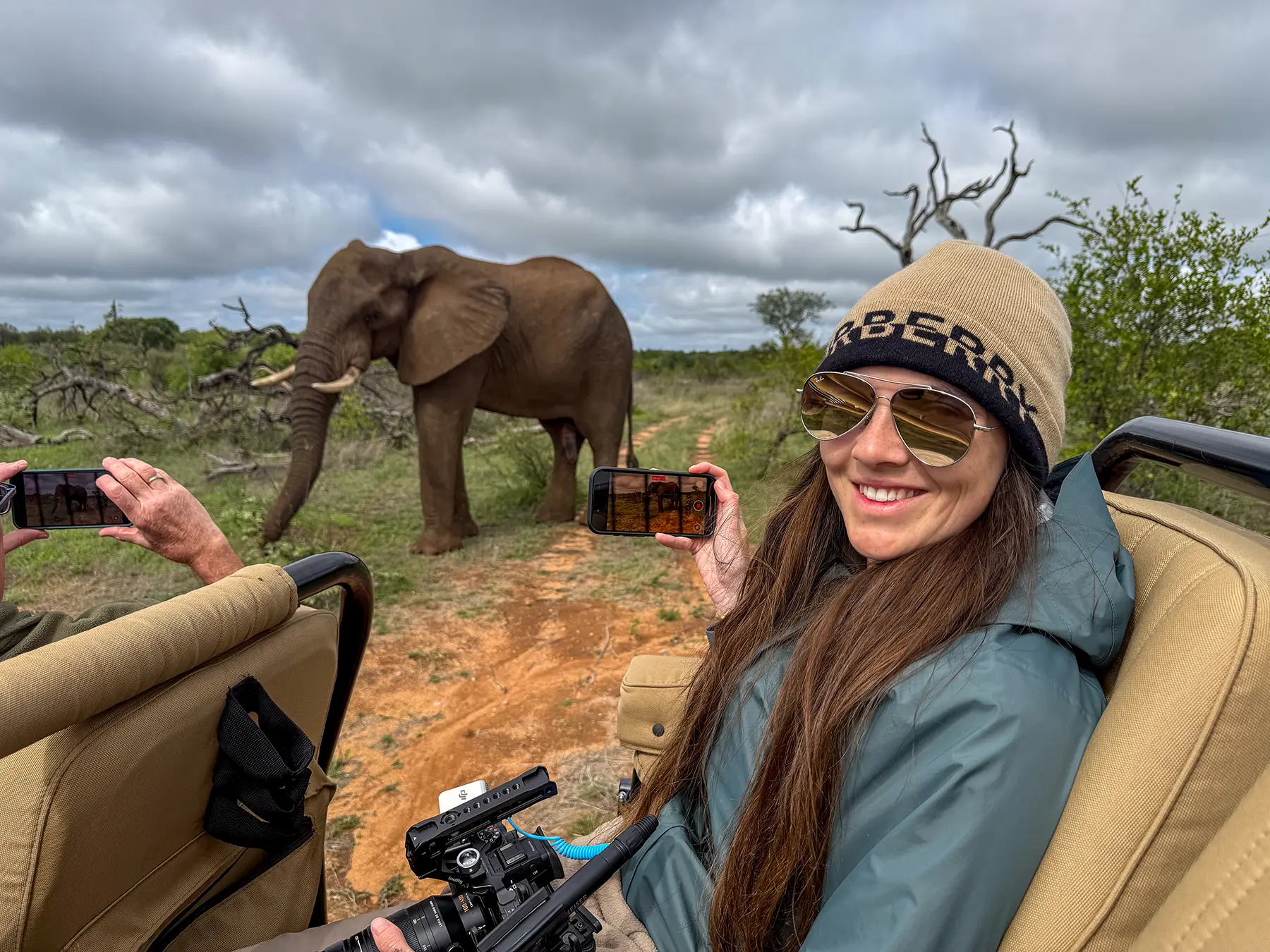 Ella McKendrick smiling and holding up her phone as a bull elephant walks past the safari vehicle on safari in Olifants West Game Reserve, South Africa
