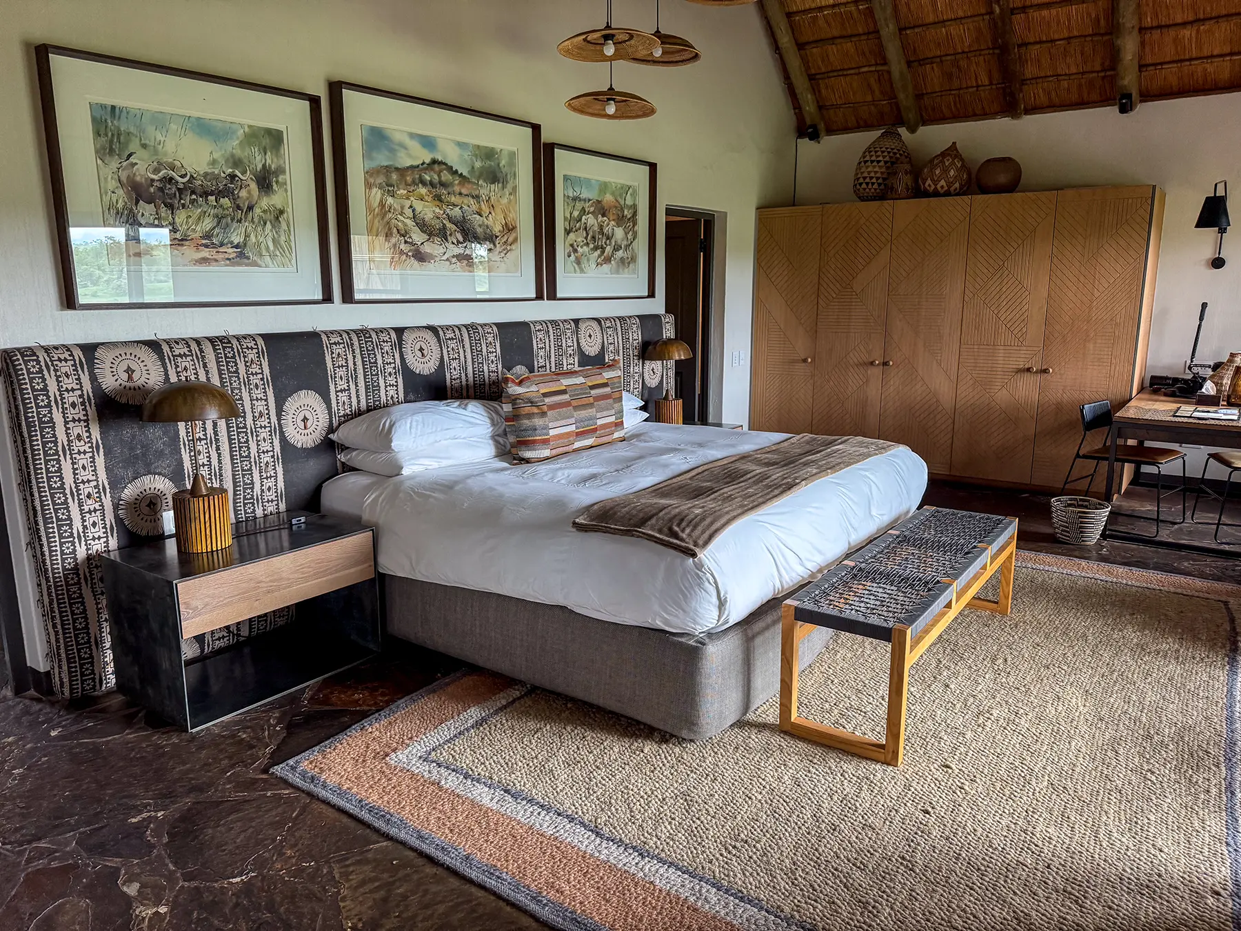 A spacious bedroom with a king-size bed, patterned African headboard, framed wildlife prints, and a thatched ceiling on safari in Sabi Sand Game Reserve, South Africa