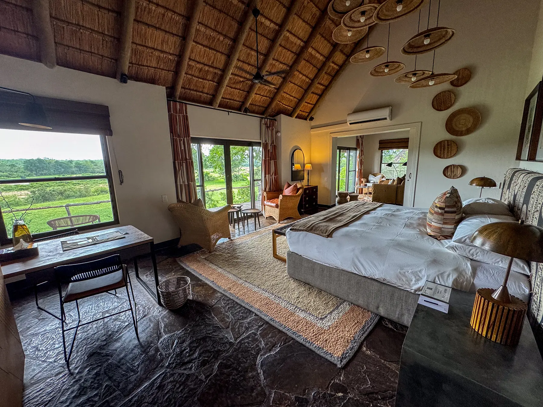 A bedroom with a king bed, writing desk, and large windows overlooking a green river floodplain, with woven basket wall decor on safari in Sabi Sand Game Reserve, South Africa
