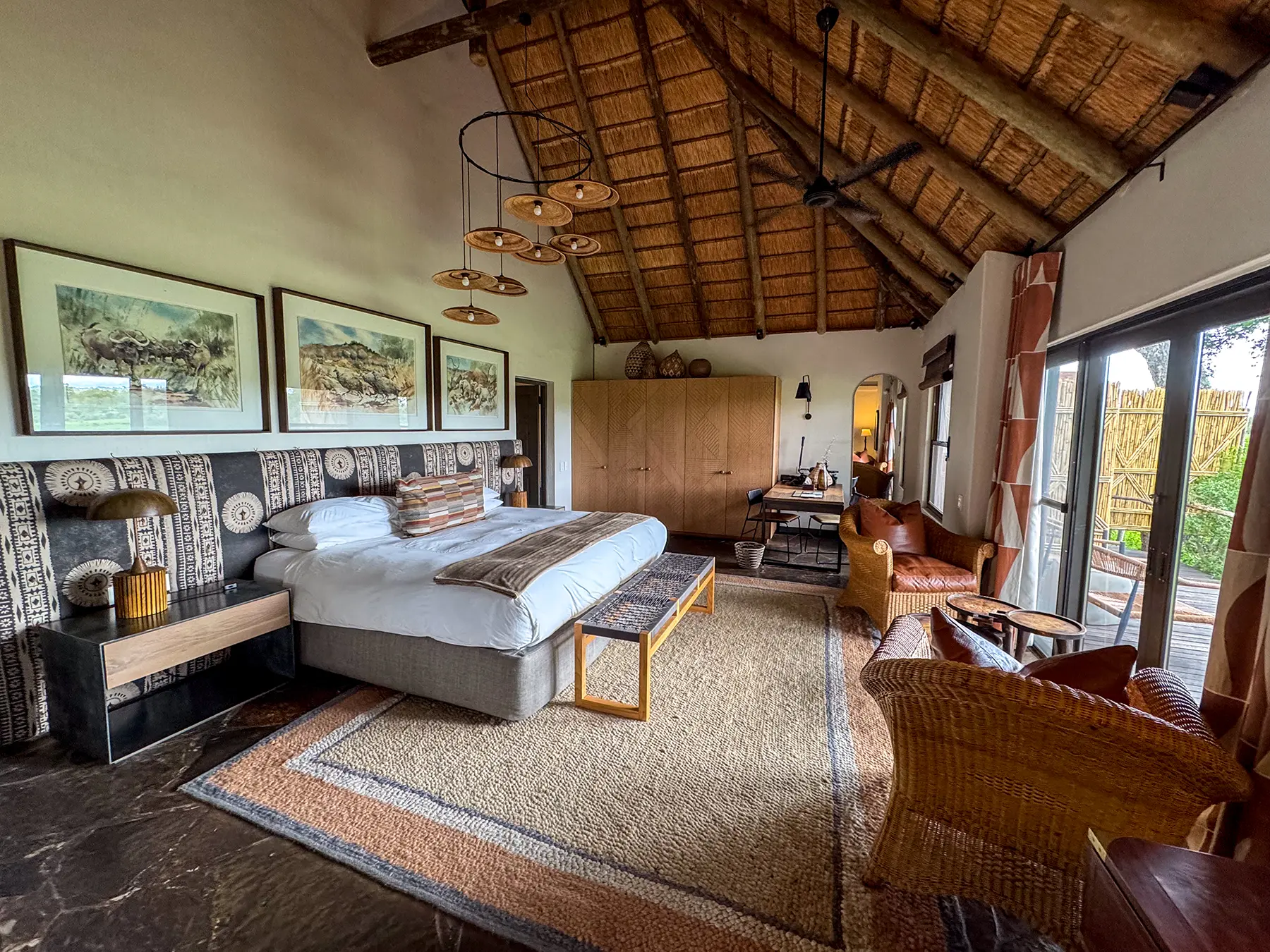 A wide-angle view of a spacious safari lodge bedroom with a king bed, rattan seating area, and pendant lights under a thatched roof on safari in Sabi Sand Game Reserve, South Africa