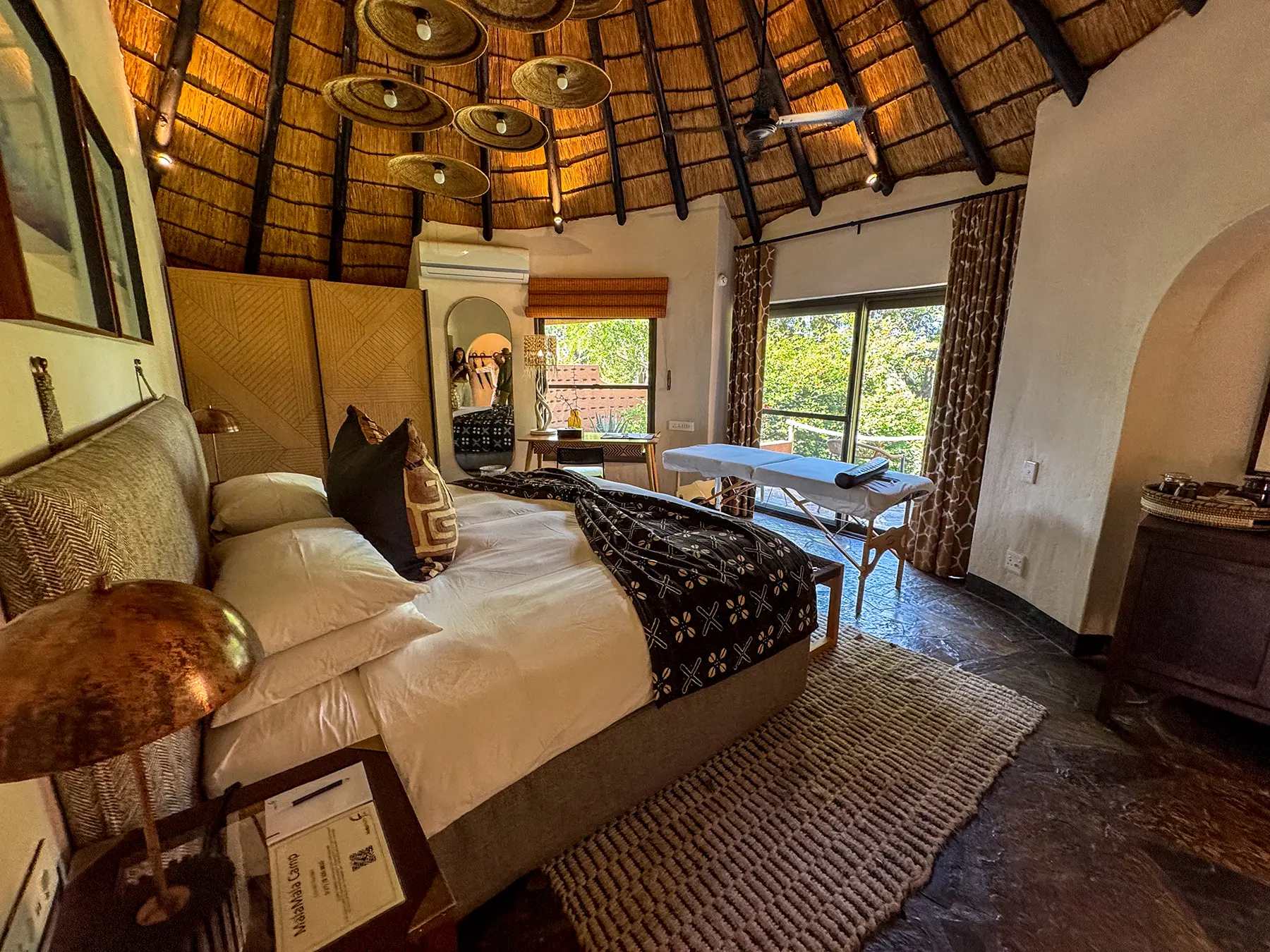 A rondavel-style bedroom with a domed thatched ceiling, king bed dressed in white linen, brass pendant lights, and a massage table on safari in the Sabi Sand Game Reserve, South Africa