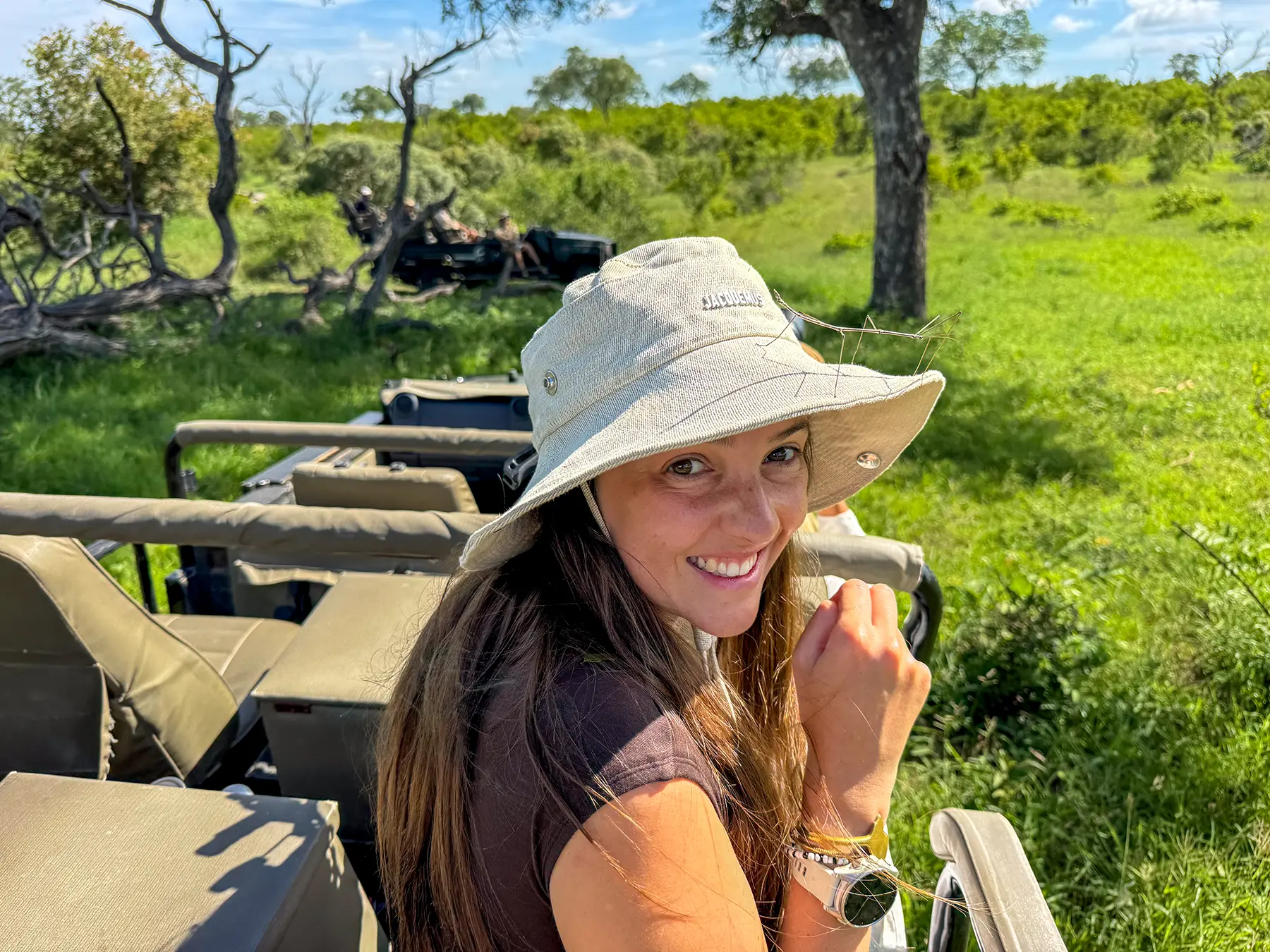 Ella McKendrick smiling in a safari hat aboard an open game drive vehicle with green bushveld behind on safari in Sabi Sand Game Reserve, South Africa