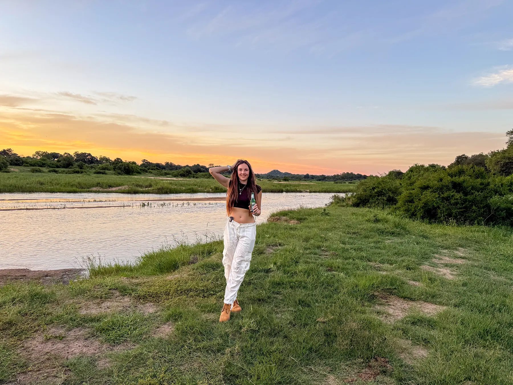 Ella McKendrick standing by a river at sunset holding a drink with pink and gold sky behind on safari in Sabi Sand Game Reserve, South Africa