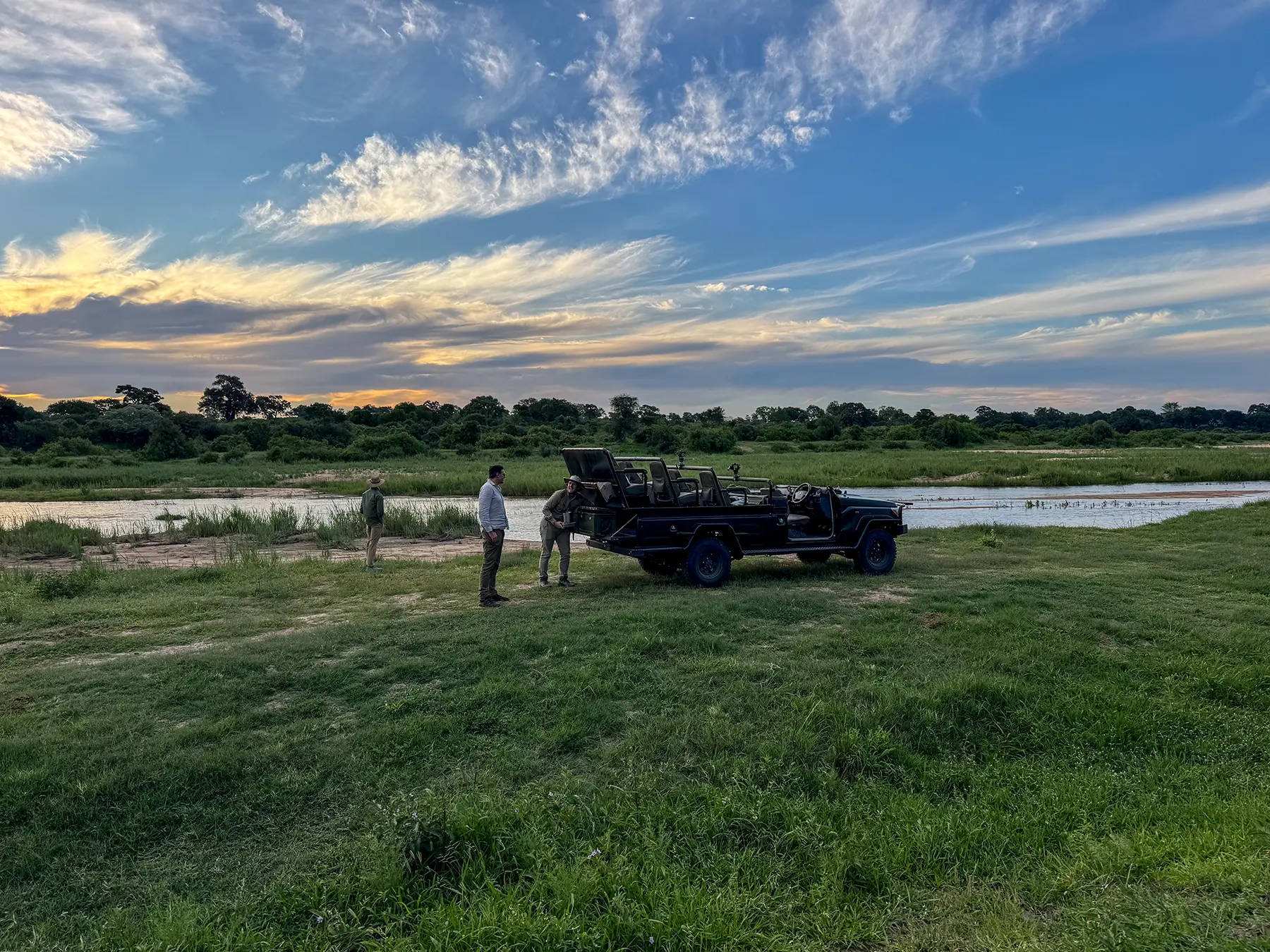 A sundowner drinks stop beside a safari vehicle on the banks of a river at sunset with a dramatic sky on safari in Sabi Sand Game Reserve, South Africa