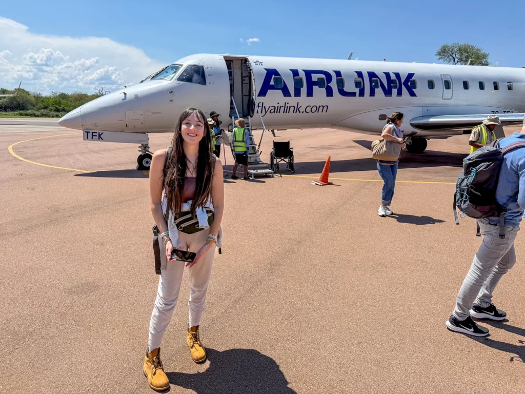 Ella McKendrick standing on the tarmac in front of an Airlink Embraer jet with passengers boarding behind her on a flight in South Africa