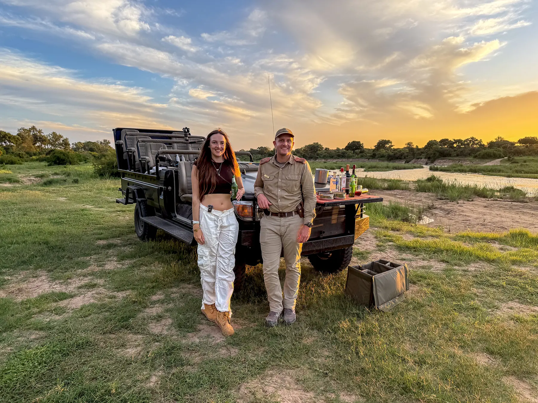Ella McKendrick with a local South African safari guide standing beside the game drive vehicle at a sunset drinks stop beside a waterhole on safari in South Africa