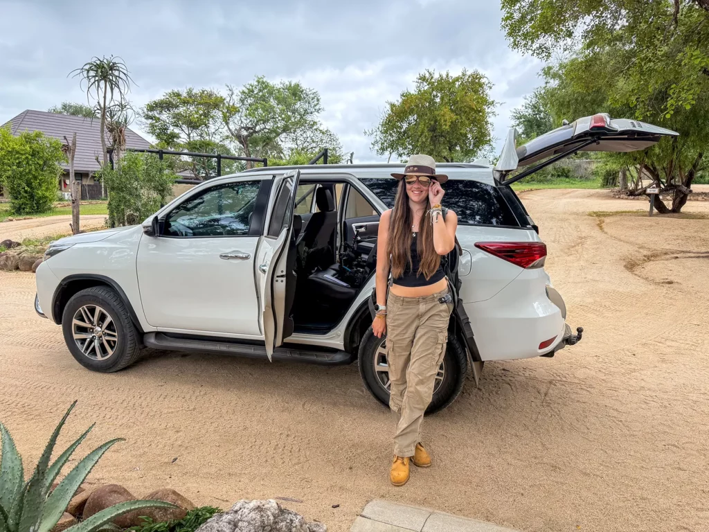 Ella McKendrick standing beside a white Toyota Fortuner with the boot open, arriving at a safari lodge in South Africa