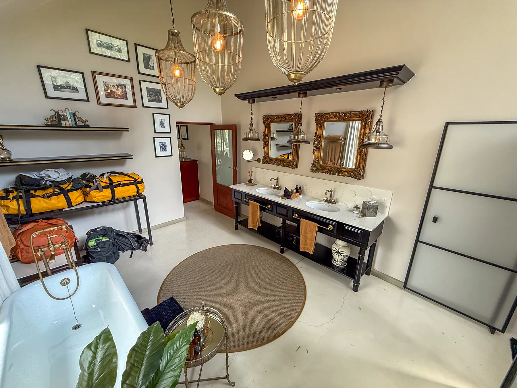 A wide-angle view of a luxury bathroom with freestanding bath, double vanity, birdcage pendant lights, gallery wall and safari luggage on safari in the Timbavati Private Nature Reserve, South Africa