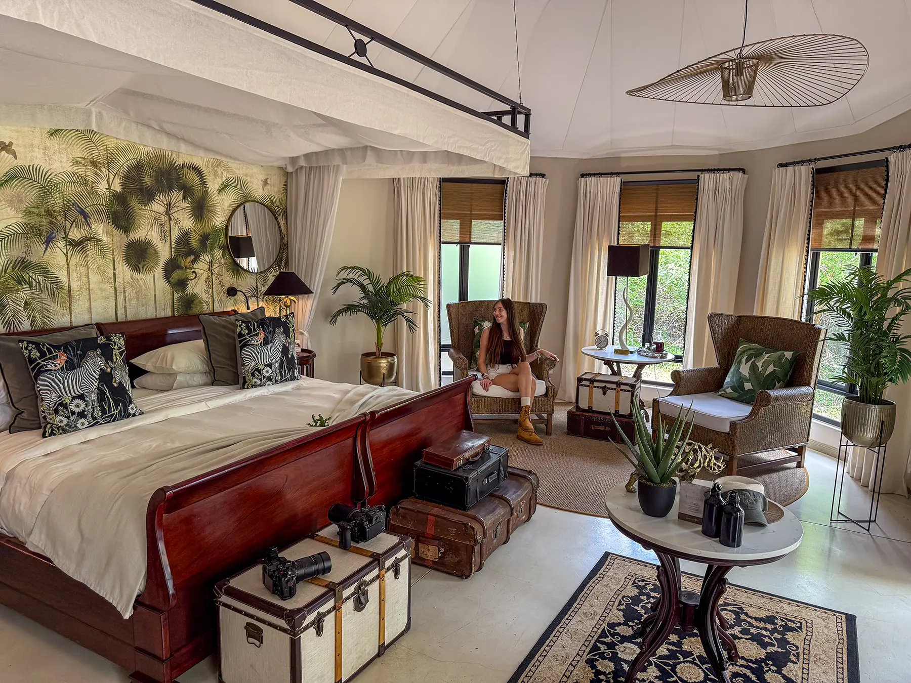 Ella McKendrick relaxing in a wingback chair in a spacious luxury suite with canopy bed, botanical wallpaper and vintage trunks on safari in the Timbavati Private Nature Reserve, South Africa