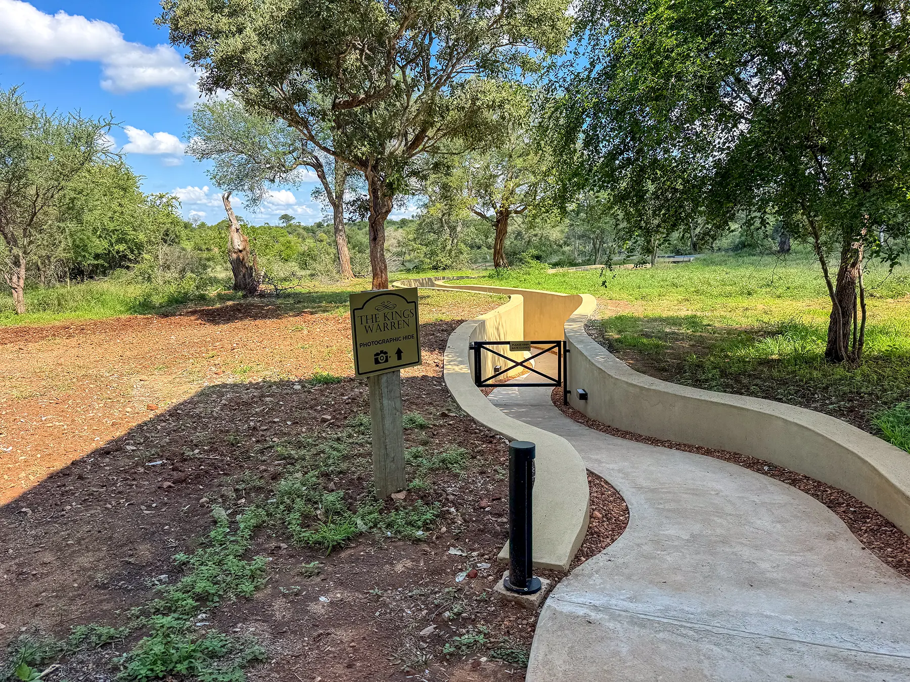 A curved pathway with a wooden signpost and gate leading to a photography hide through green bushveld on safari in the Timbavati Private Nature Reserve, South Africa