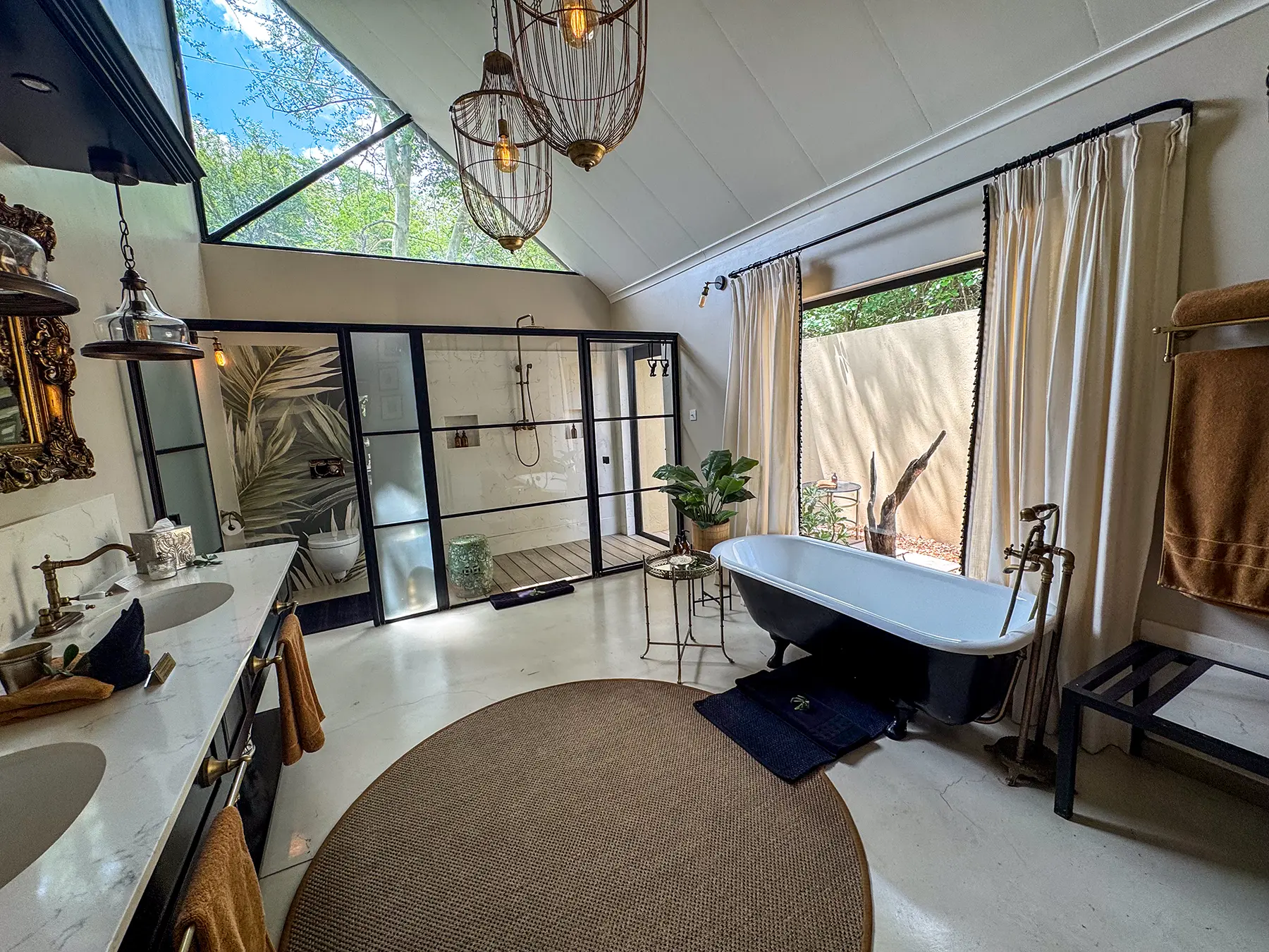 A spacious bathroom with freestanding clawfoot bath, glass-walled rain shower, birdcage pendant lights and angled skylight windows on safari in the Timbavati Private Nature Reserve, South Africa