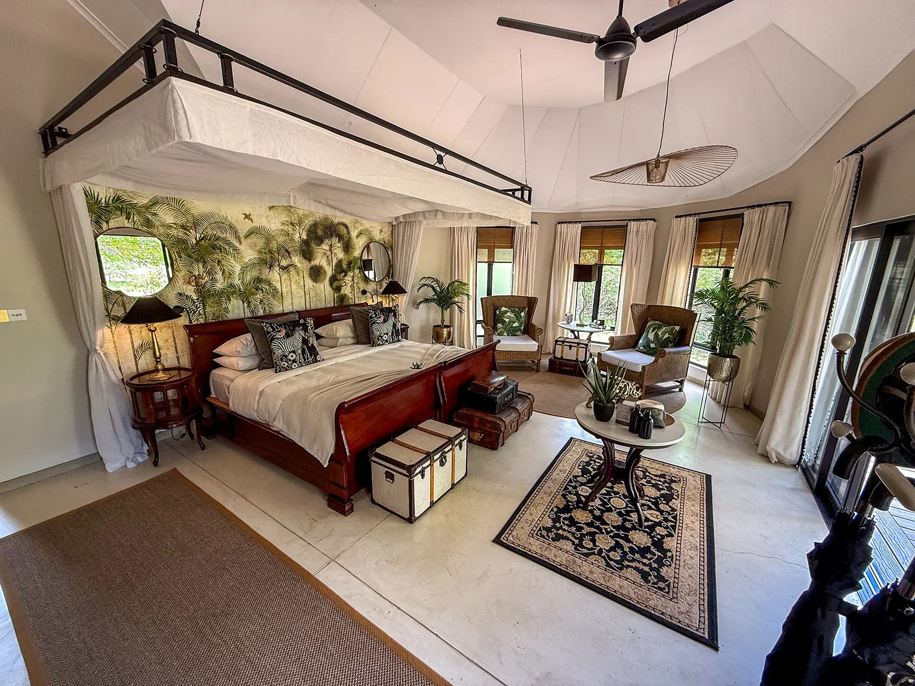 A luxury suite bedroom with four-poster canopy bed, botanical palm wallpaper, vintage trunks, Persian rug and wingback seating area on safari in the Timbavati Private Nature Reserve, South Africa
