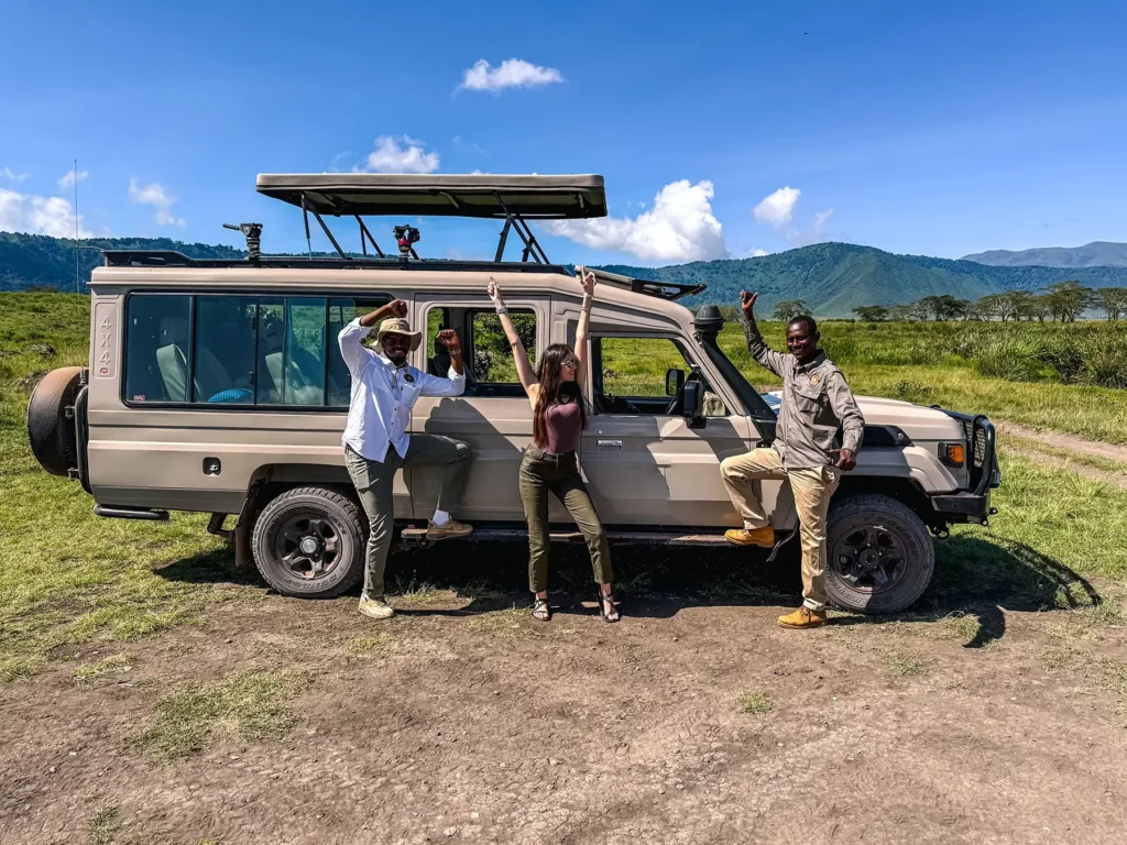 Ella McKendrick celebrating with two guides beside a safari vehicle in Ngorongoro Crater, Tanzania