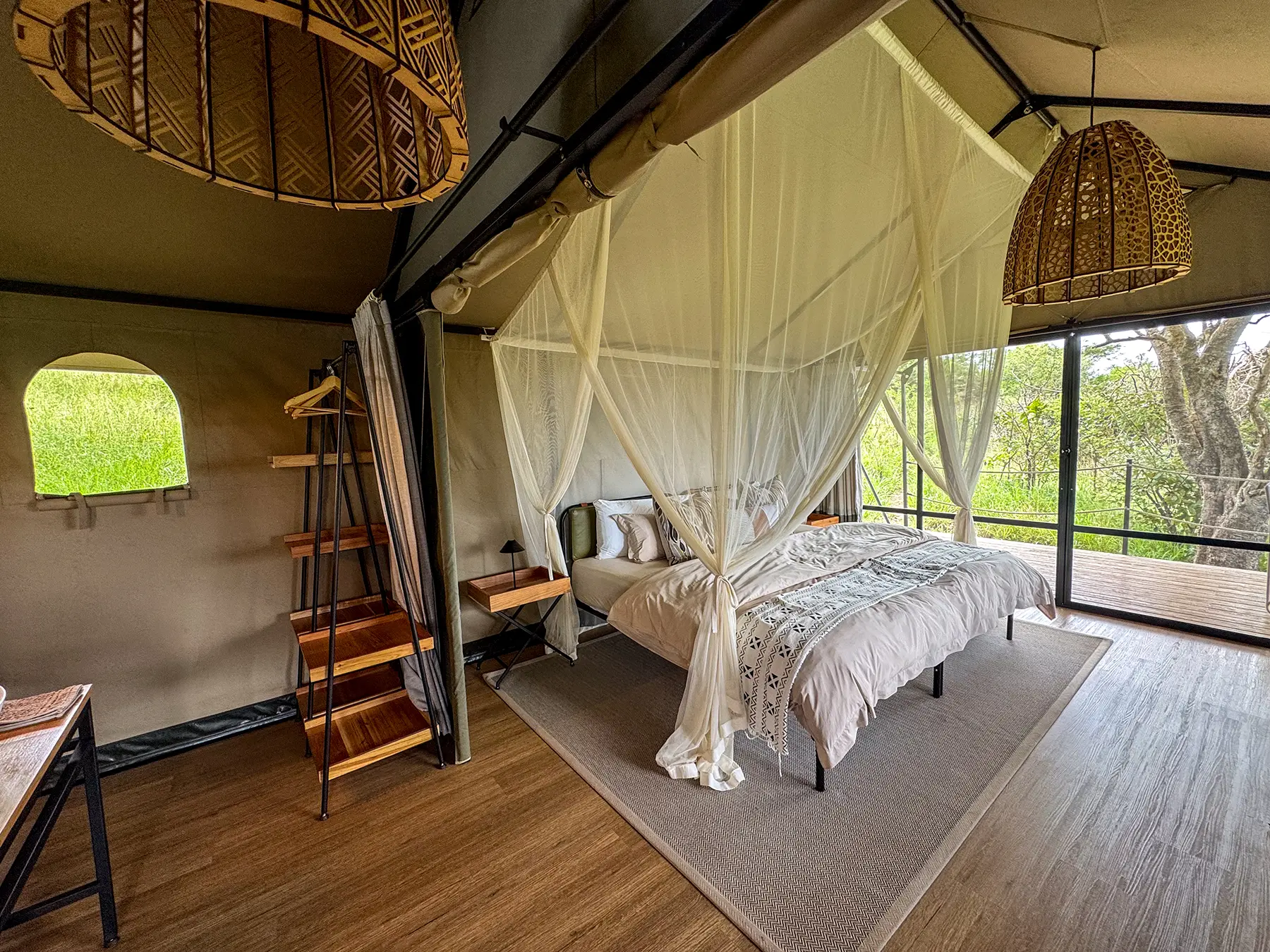 Four-poster bed draped in mosquito netting beside floor-to-ceiling glass overlooking greenery at Karenge Bush Camp in Akagera National Park, Rwanda