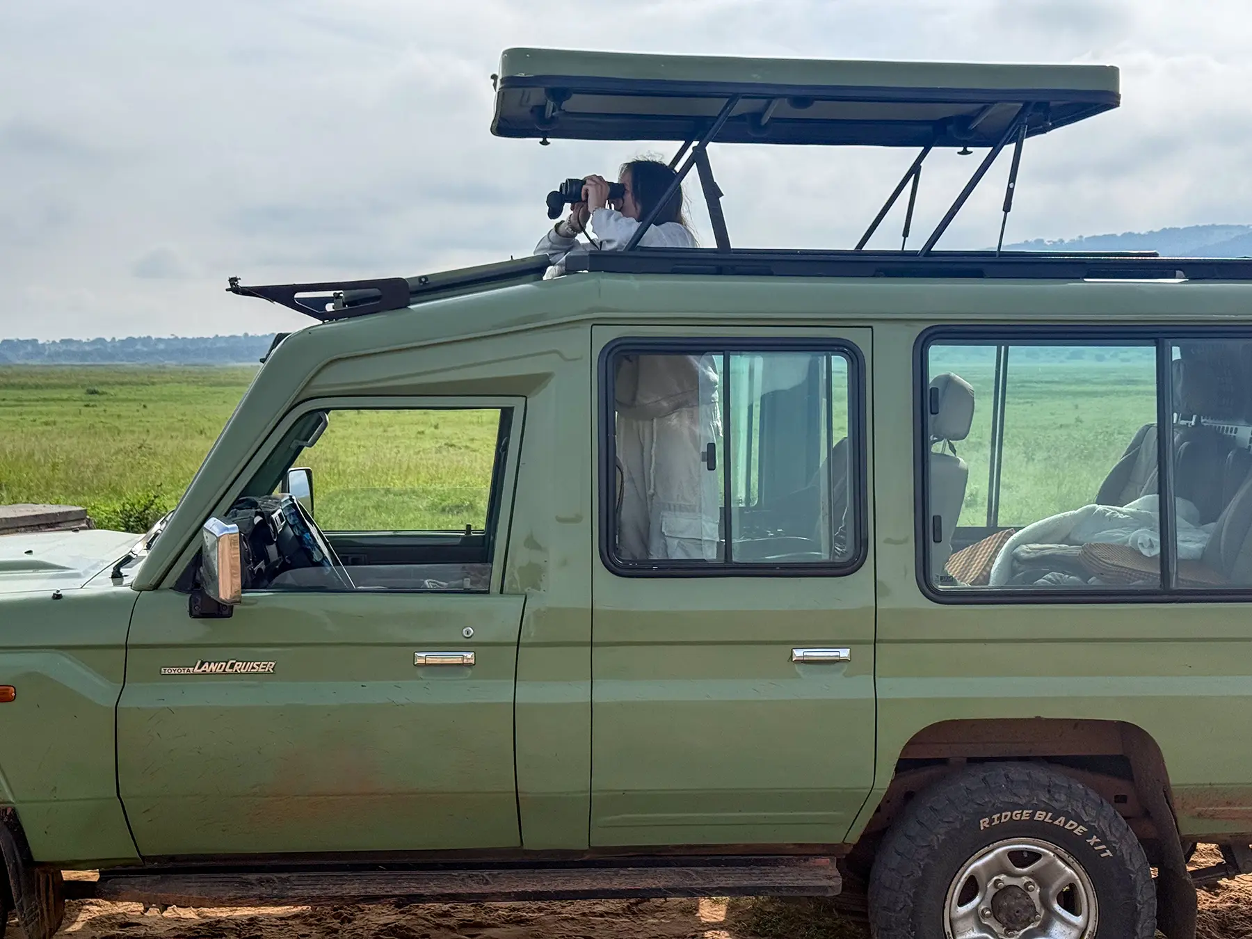 Ella McKendrick scanning with binoculars from raised roof of green Land Cruiser on safari in Akagera National Park, Rwanda