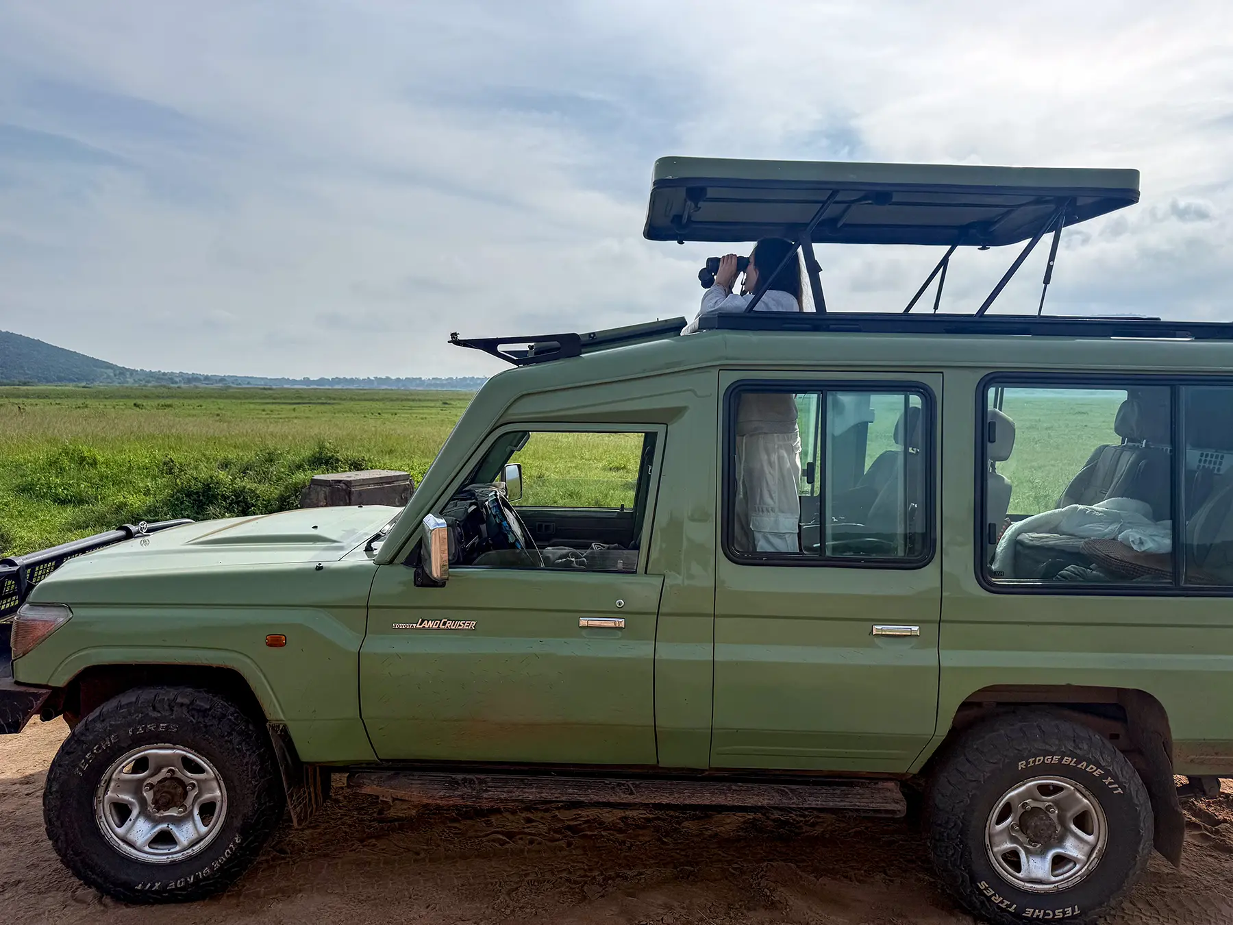Ella McKendrick using binoculars from the pop-top of a green Land Cruiser on safari in Akagera National Park, Rwanda