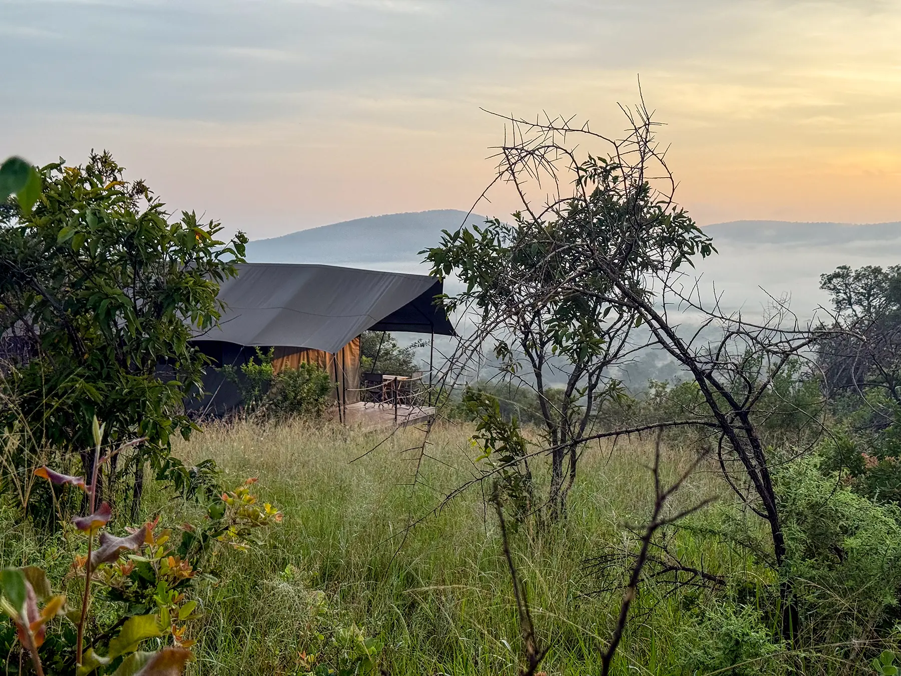 Safari tent on grassy hillside catches first light over misty valley at Karenge Bush Camp in Akagera National Park, Rwanda