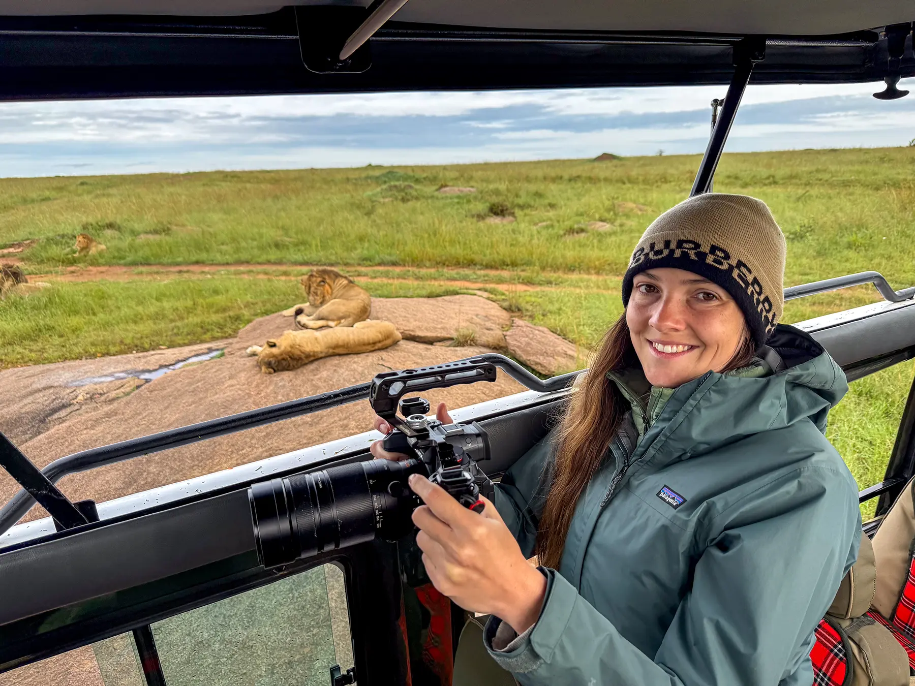Ella McKendrick holding a telephoto camera lens inside a vehicle, with a resting lion behind on safari