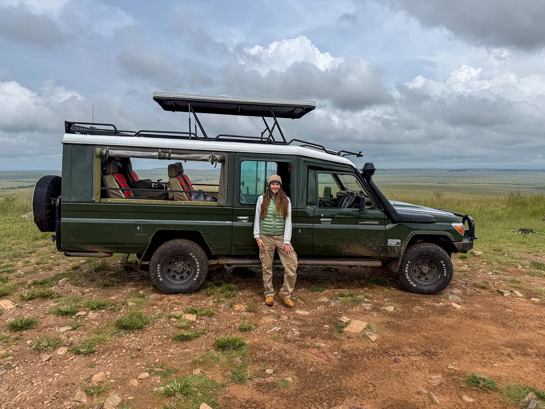 Ella McKendrick stands beside a green Land Cruiser with a pop-top roof on a stony ridge on safari