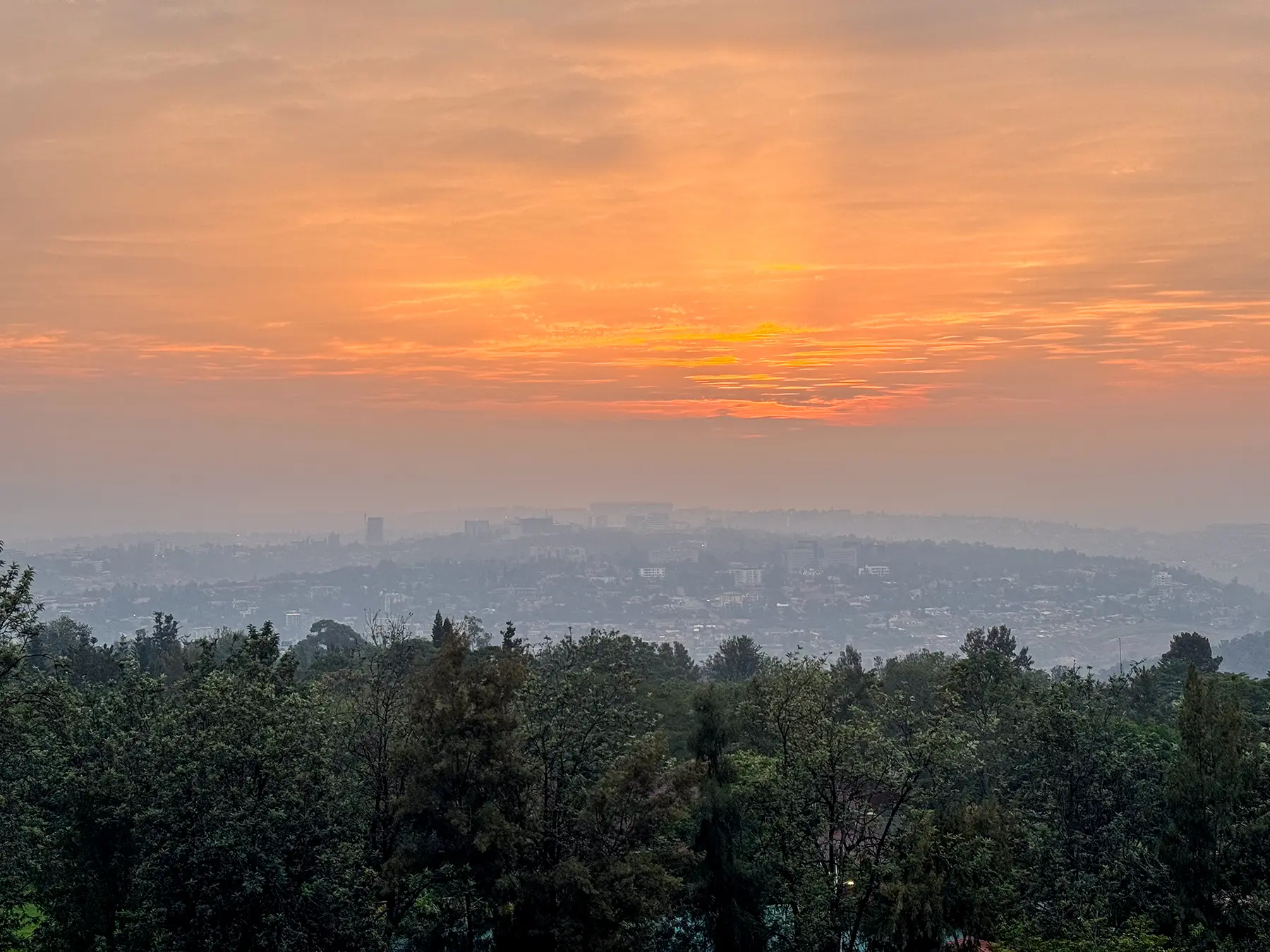 Glowing orange sky over hazy city skyline and treetops at hilltop viewpoint in Kigali, Rwanda