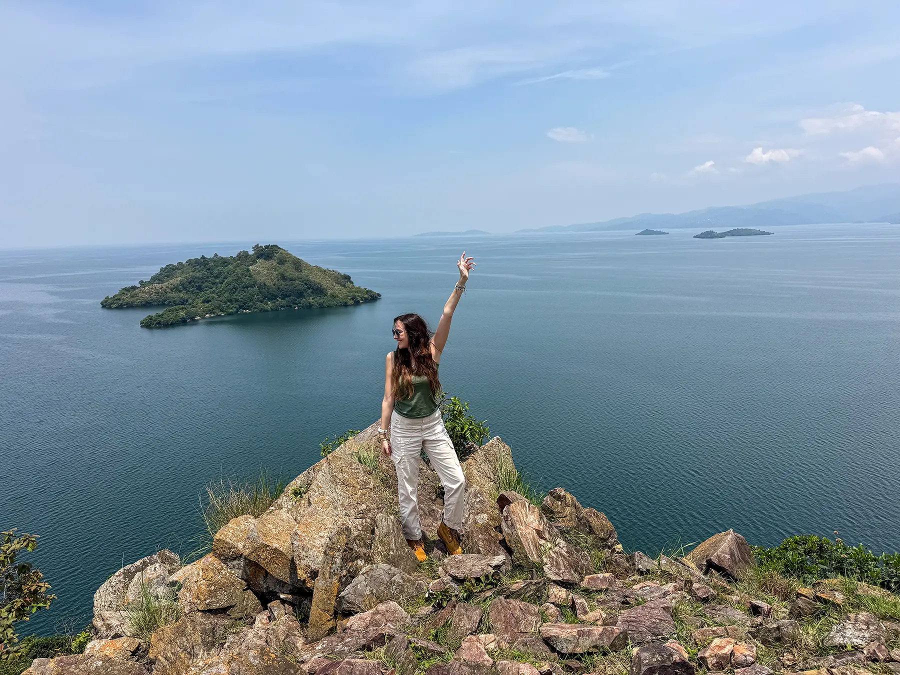 Ella McKendrick on rocky outcrop with arm raised above an island view at Lake Kivu, Rwanda
