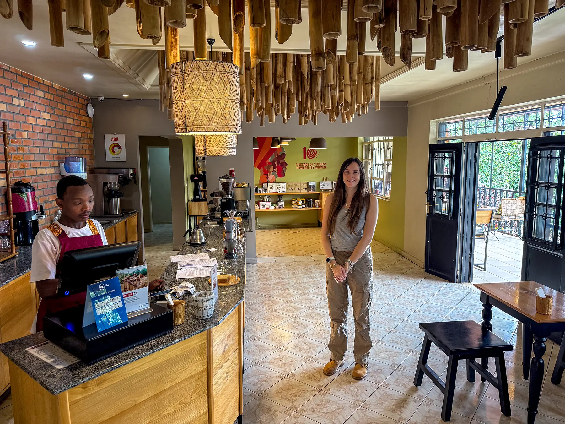 Ella McKendrick standing beside the bamboo-ceilinged counter with a barista at Question Coffee in Kigali, Rwanda