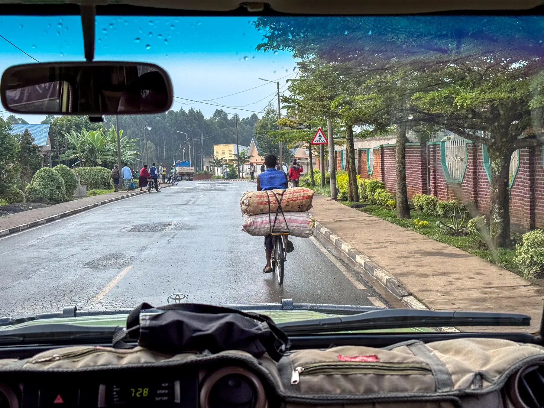 Cyclist carrying heavy load on rural road viewed through vehicle windscreen at journey route, Rwanda