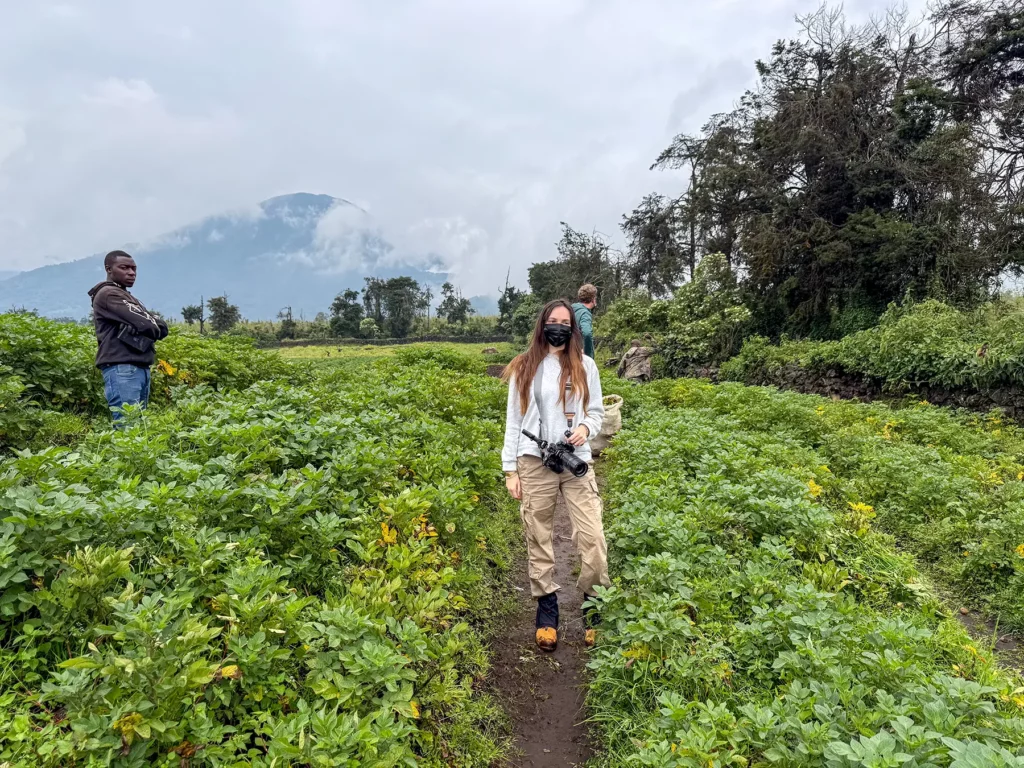 Ella McKendrick on narrow path through green crop fields with face mask and camera at Volcanoes National Park, Rwanda
