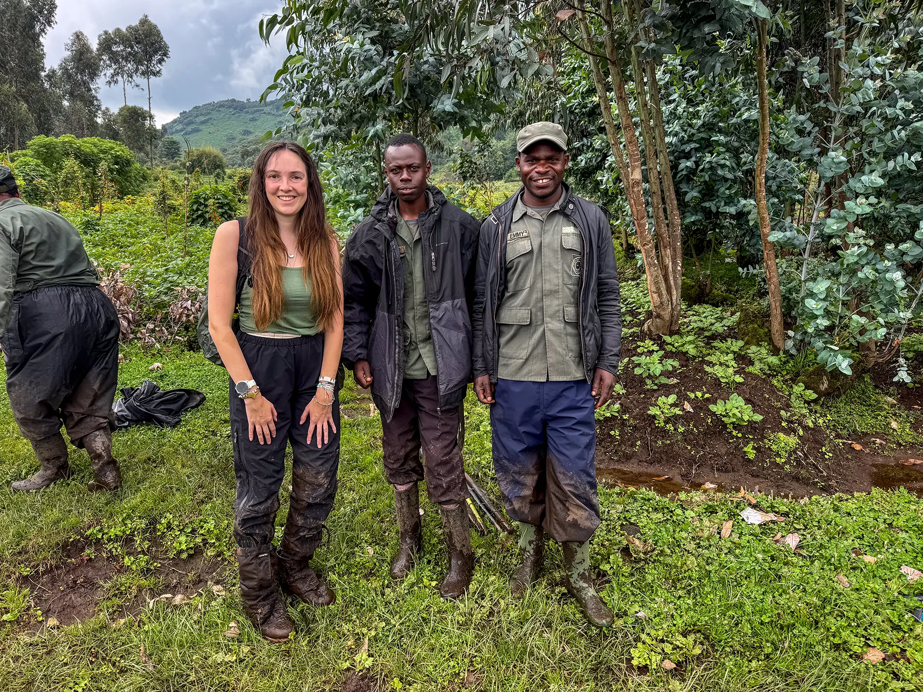 Ella McKendrick smiling between two Rwandan rangers in green uniforms in a grassy clearing at Volcanoes National Park, Rwanda