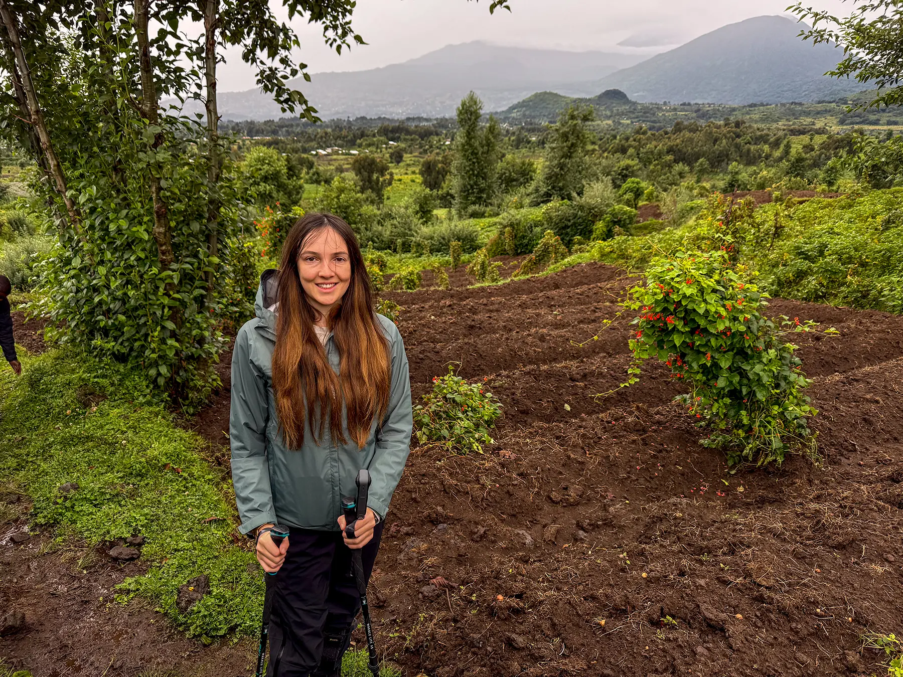 Ella McKendrick with trekking poles on tilled earth before volcanic mountains under cloudy sky at the briefing area in Volcanoes National Park, Rwanda