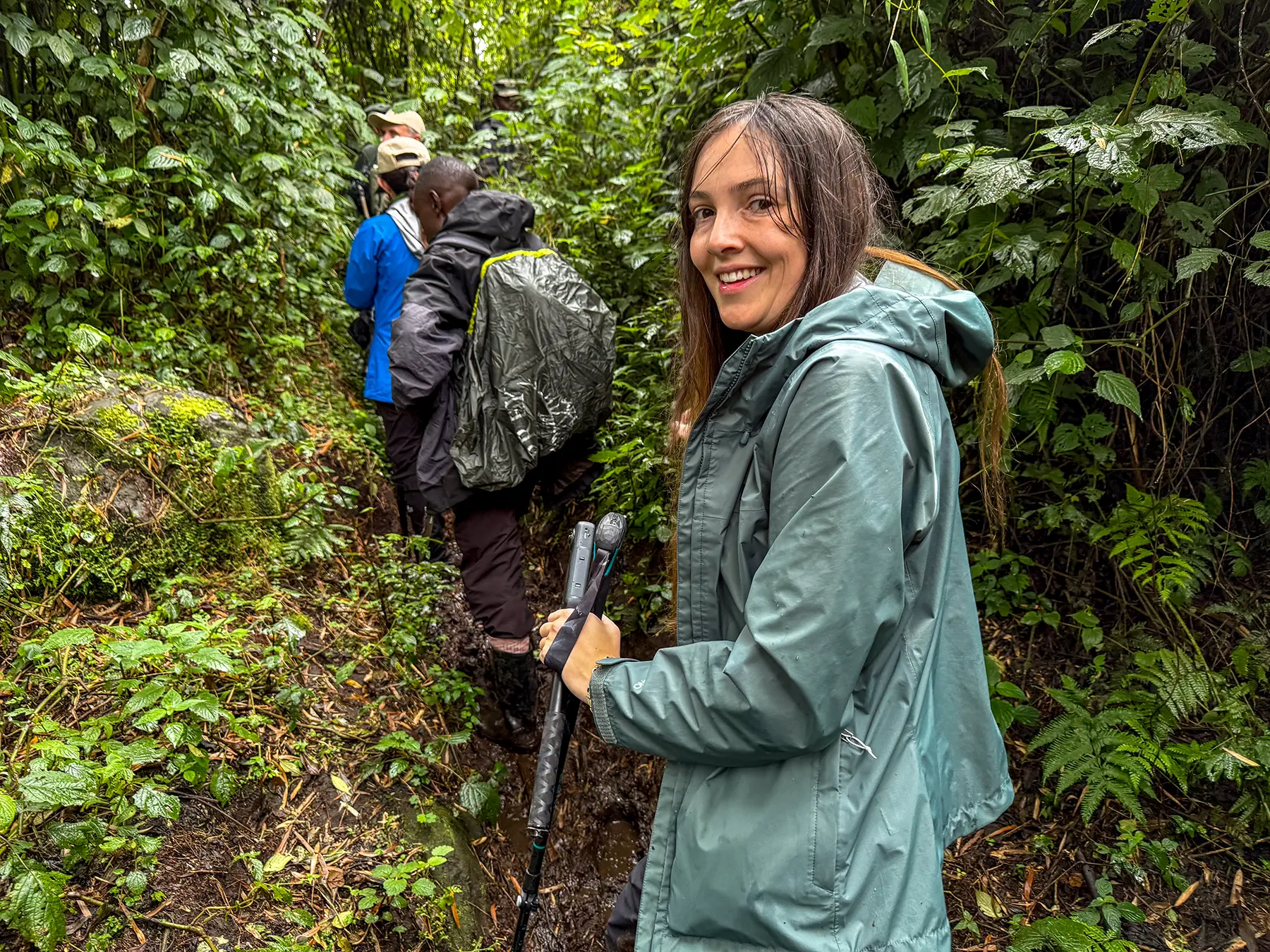 Ella McKendrick smiling on a forest trail with porters and guides ahead at the lower slopes in Volcanoes National Park, Rwanda