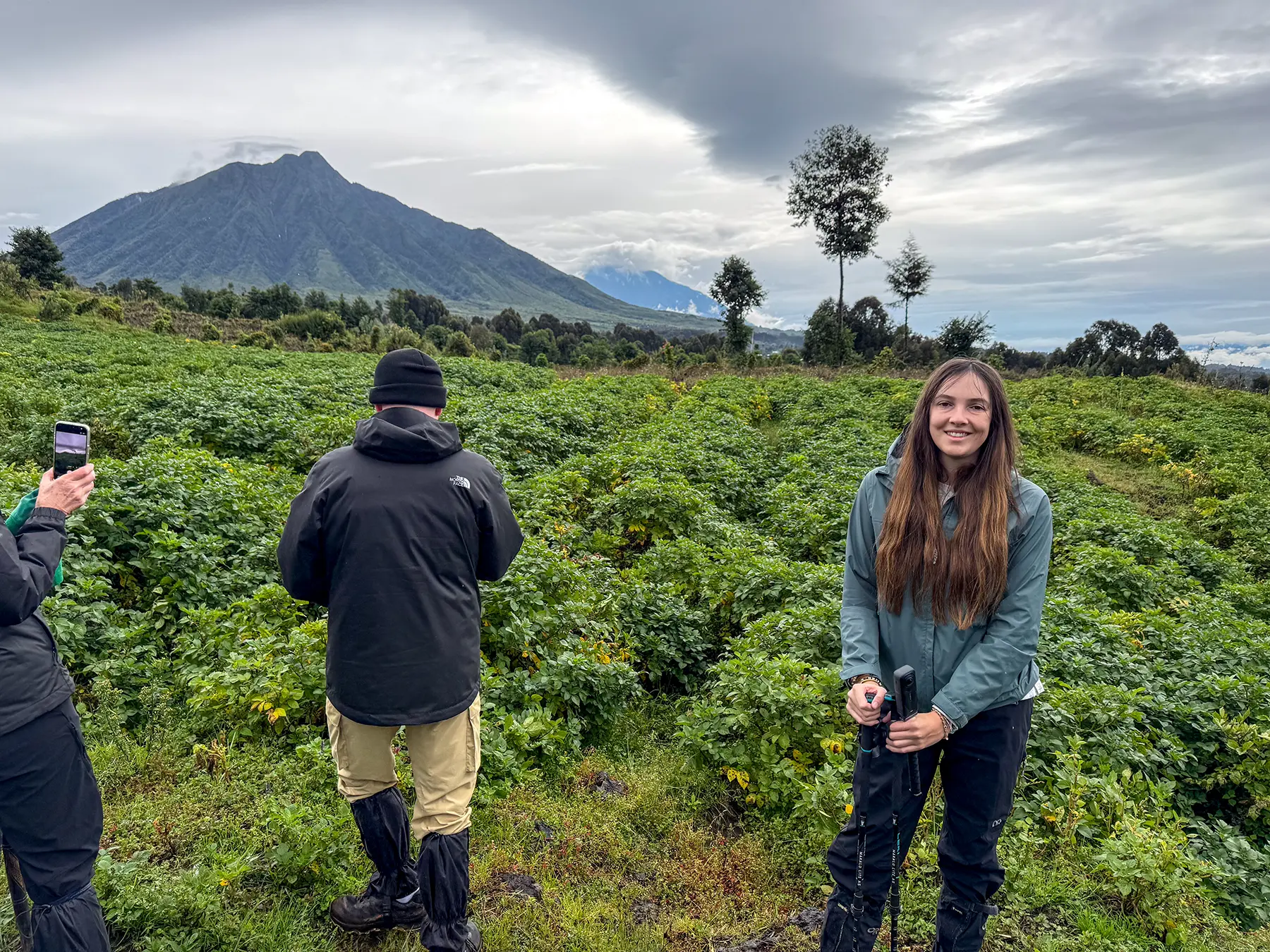 Ella McKendrick smiling beside two figures with camera and distant volcano across green crops at the briefing area in Volcanoes National Park, Rwanda