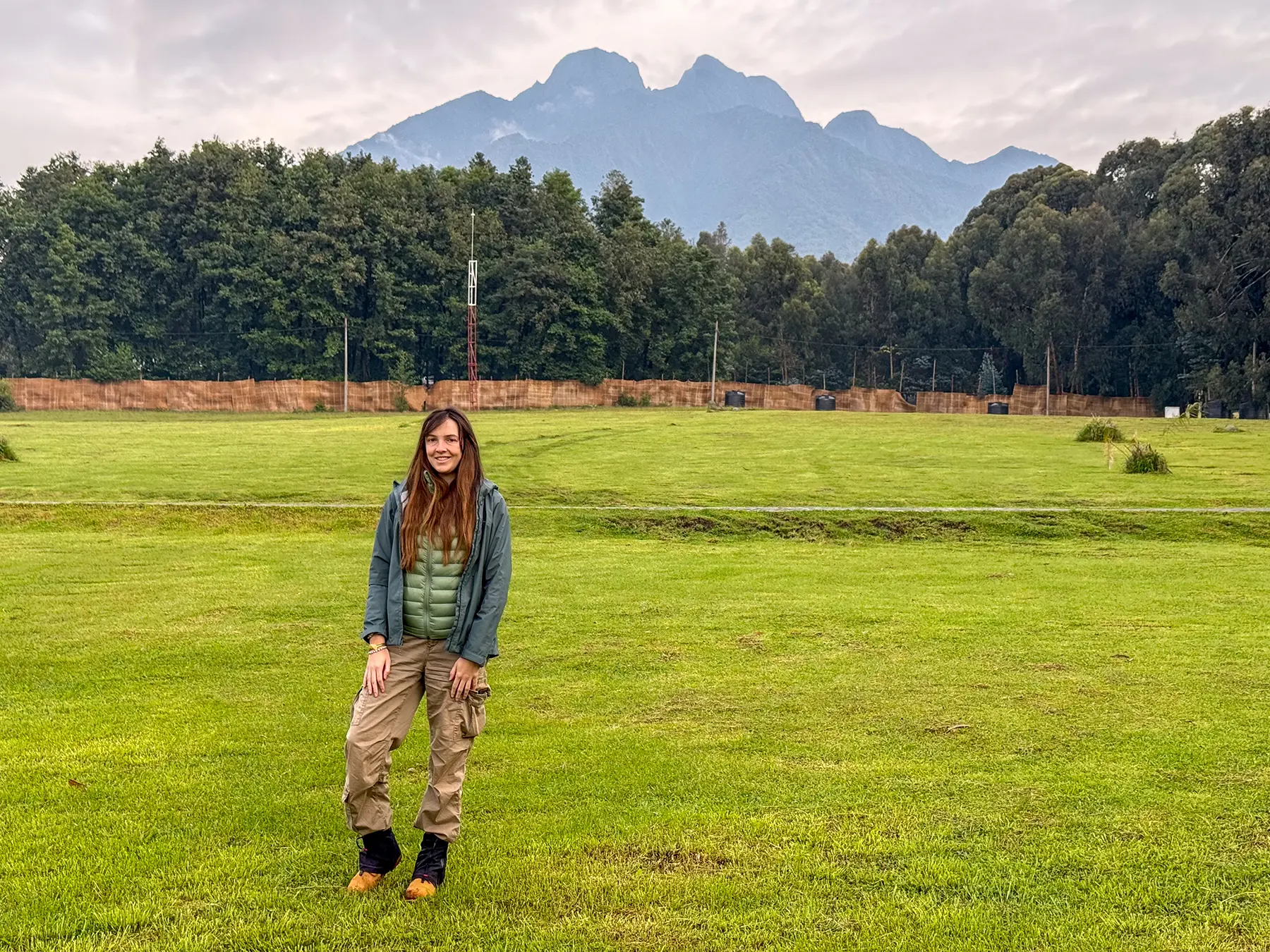 Ella McKendrick standing in a grassy field with the Virunga volcanoes rising behind at Kinigi sector in Volcanoes National Park, Rwanda