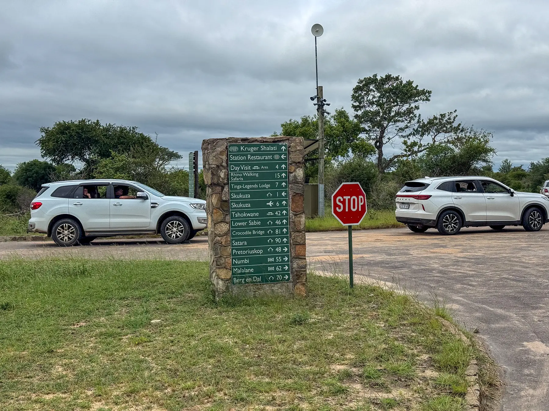 Stone directional signpost listing destinations and distances with cars at a road junction at Skukuza in Kruger National Park, South Africa