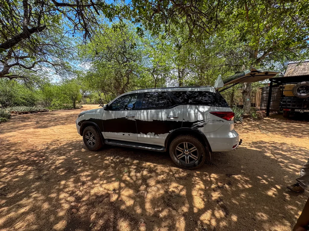 White SUV with raised tailgate parked on sandy ground at a shaded parking area in South Africa