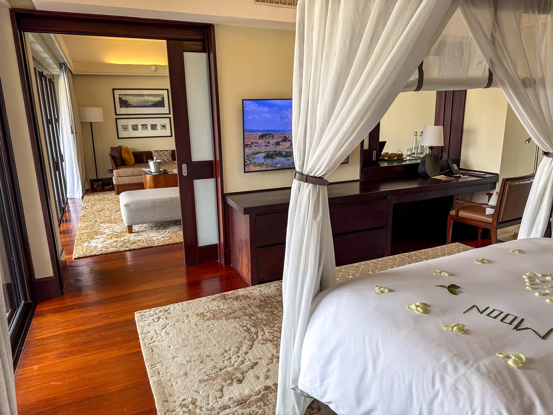 Four-poster bed scattered with petals and a desk with the suite living room beyond at Four Seasons Safari Lodge Serengeti in Serengeti National Park, Tanzania