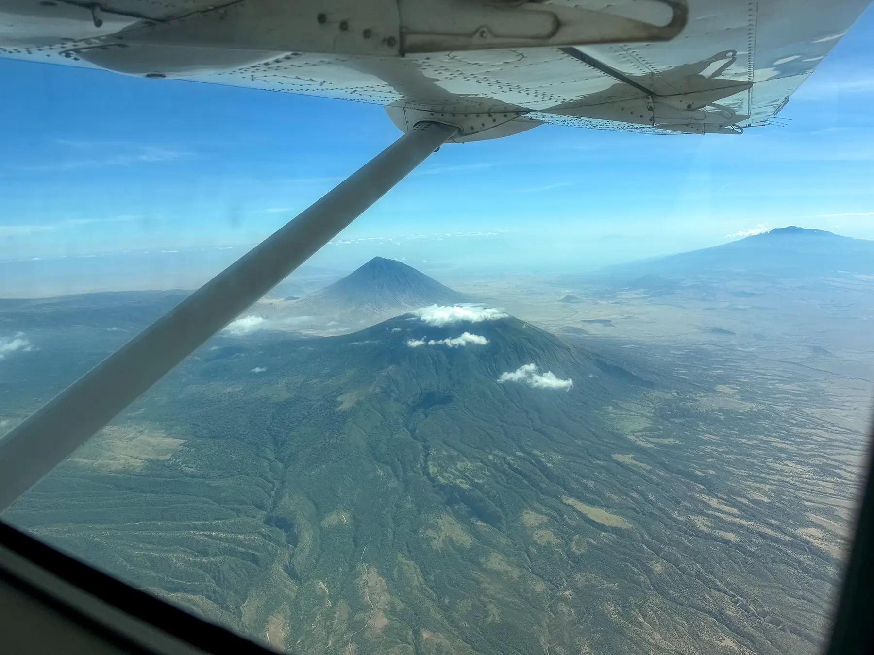 Cessna Caravan wing strut framing a sharp volcanic cone with distant peaks beyond at Ol Doinyo Lengai, Tanzania