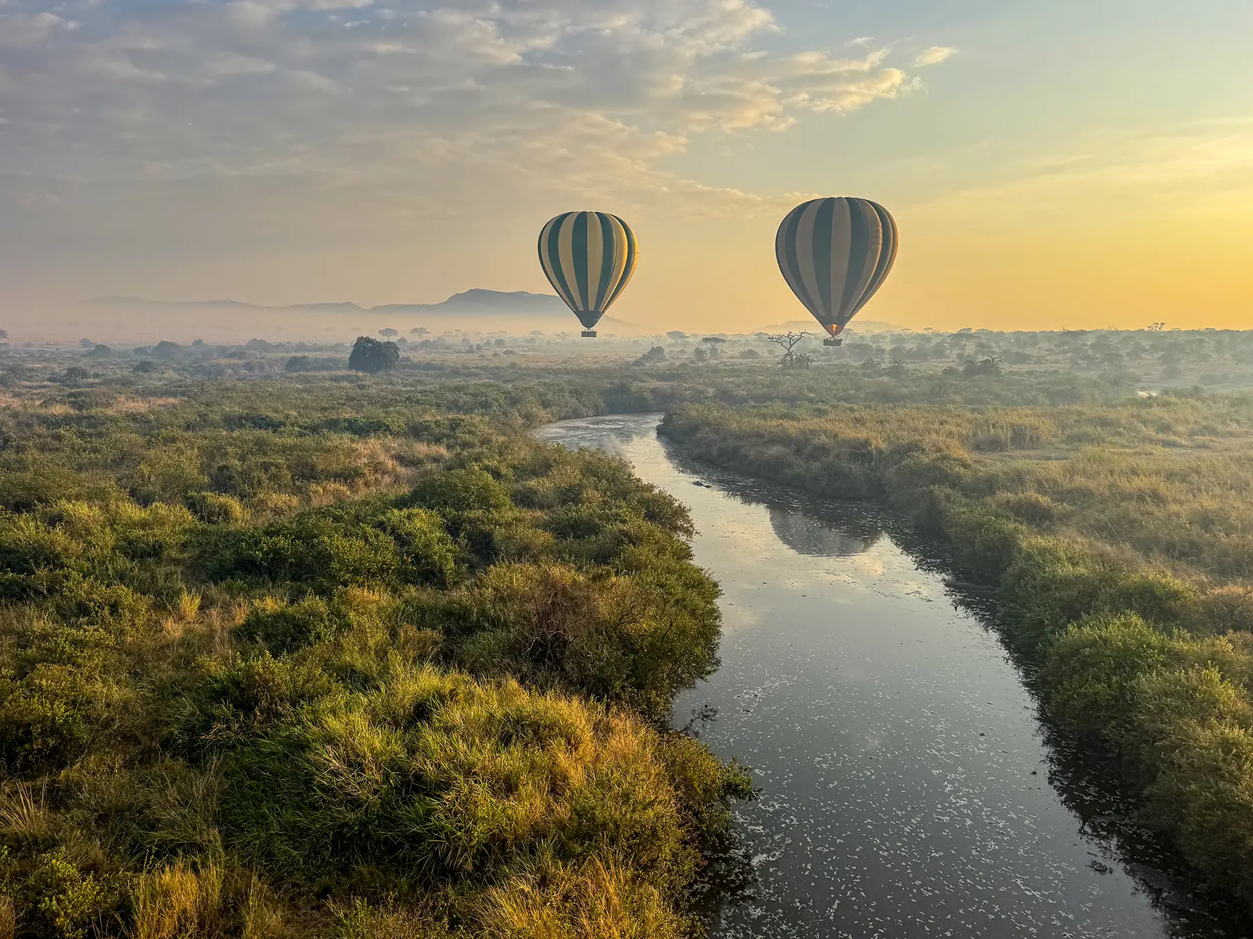 Two hot air balloons above a meandering river and green scrub in early light on safari in Serengeti National Park, Tanzania