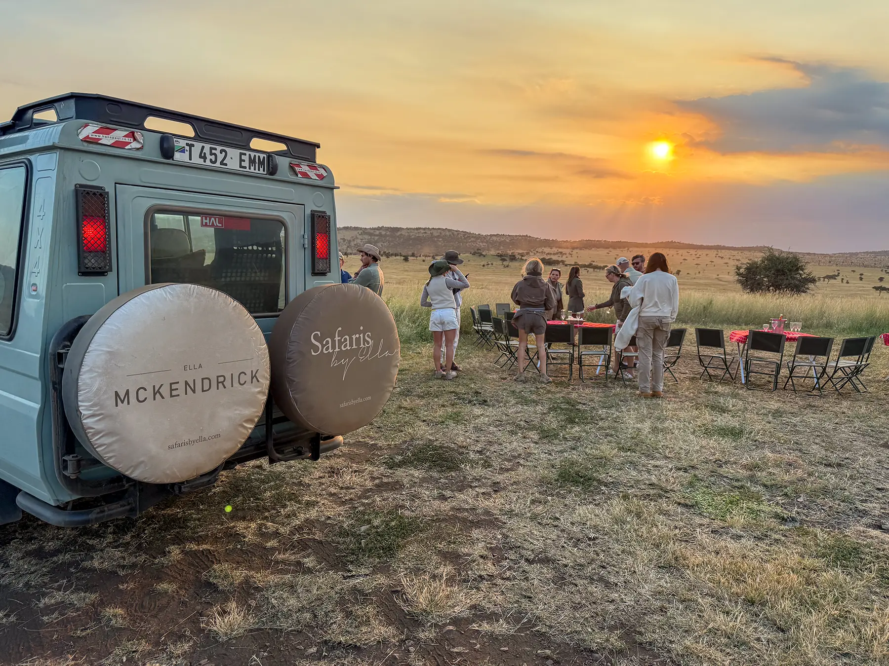 Ella McKendrick with guests at sundowner drinks around folding tables beside a Land Cruiser on safari in Serengeti National Park, Tanzania