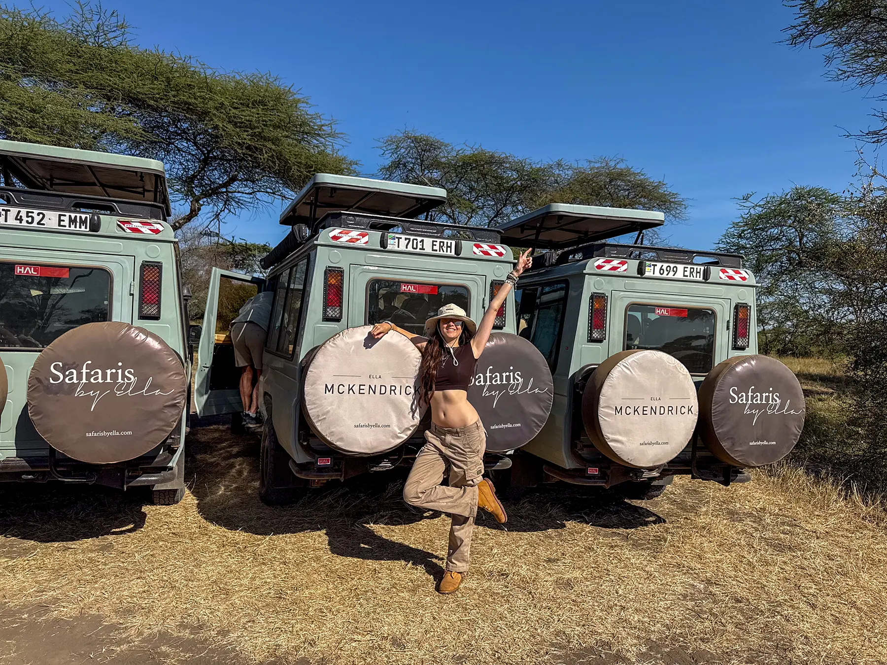 Ella McKendrick standing beside parked Land Cruisers with spare wheel covers on safari in Serengeti National Park, Tanzania