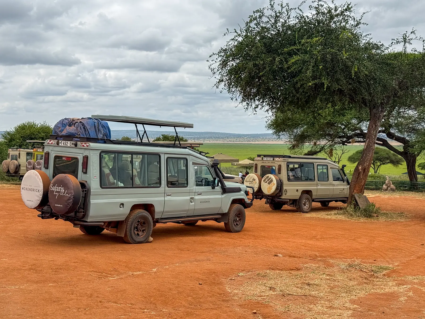 Safari vehicles parked on red earth beside picnic shelters above green plains on safari in Tarangire National Park, Tanzania